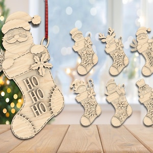 Puede incluir: Adornos de calcetín navideño de madera con Papá Noel, muñecos de nieve, renos y cascanueces. El calcetín de Papá Noel tiene el texto "HO HO HO". Cada adorno está hecho de madera de color claro y tiene una cinta roja para colgar.