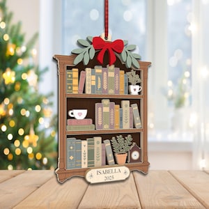 May include: Wooden Christmas tree ornament shaped like a bookshelf, with a red bow, green leaves, and a red ribbon. The bookshelf is filled with books, a teacup, and a small plant. The ornament has the name "Isabella" and the year "2025".