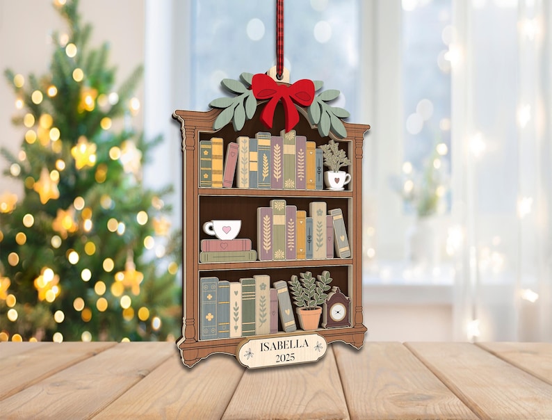 May include: Wooden Christmas tree ornament shaped like a bookshelf, with a red bow, green leaves, and a red ribbon. The bookshelf is filled with books, a teacup, and a small plant. The ornament has the name "Isabella" and the year "2025".