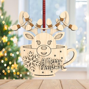 May include: Wooden Christmas ornament depicting a reindeer head in a teacup shape. The ornament has a snowflake and the text "A Merry Christmas". The reindeer has antlers with light bulb accents. A red and black plaid ribbon is attached for hanging.