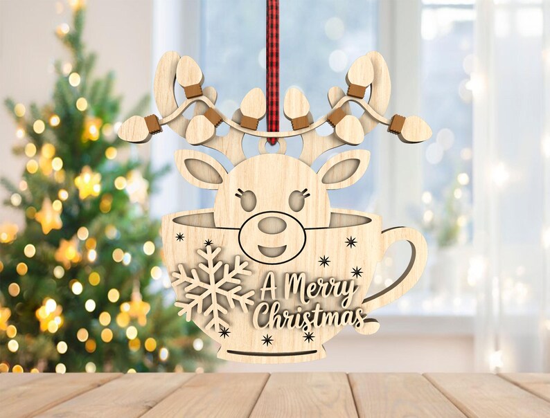 May include: Wooden Christmas ornament depicting a reindeer head in a teacup shape. The ornament has a snowflake and the text "A Merry Christmas". The reindeer has antlers with light bulb accents. A red and black plaid ribbon is attached for hanging.