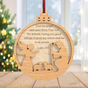 May include: Wooden Christmas ornament with a circular design. The ornament features an angel, a shepherd, and sheep, with the text "And the angel said unto them, Fear not...Luke 2:10." A red and black plaid ribbon is attached for hanging.