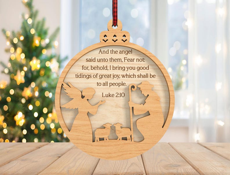 May include: Wooden Christmas ornament with a circular design. The ornament features an angel, a shepherd, and sheep, with the text "And the angel said unto them, Fear not...Luke 2:10." A red and black plaid ribbon is attached for hanging.