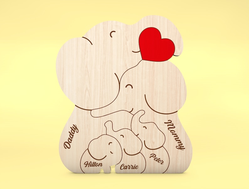 Op de afbeelding: Houten familie-olifantvormige decoratie met een rood hart. Het ontwerp bevat de woorden "Daddy" en "Mommy", samen met de namen "Hilton", "Carrie" en "Peter". De olifantenfamilie is gemaakt van lichtgekleurd hout.