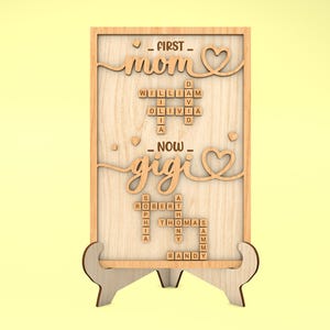 May include: Wooden sign with the words "FIRST - mom" and "NOW - gigi" in a cursive font. The words are surrounded by hearts. The sign also features a crossword puzzle with the names "William", "Olivia", "Robert", "Sophia", "Thomas", "Randy", and "Sammy".