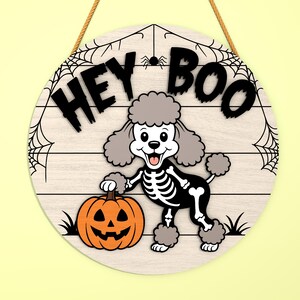 Może przedstawiać: Okrągły drewniany znak na Halloween z napisem "HEY BOO". Kresk&oacute;wkowy pudel w kostiumie szkieletu trzyma pomarańczową dynię. Znak ma wz&oacute;r pajęczyny i jest zawieszony na brązowej linie.