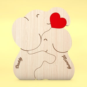 Op de afbeelding: Houten puzzel in de vorm van een olifant met een rood hart en de woorden "Daddy" en "Mommy". De puzzel is gemaakt van lichtgekleurd hout met een gladde afwerking. Het ontwerp toont twee olifanten die elkaar omarmen, met een hart ertussen.