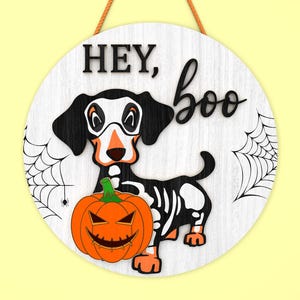 Może przedstawiać: Okrągły drewniany znak na Halloween z motywem szkieletu jamnika, pomarańczową dynią i napisem "HEY, boo". Znak ma białe tło z czarnymi pajęczynami i brązowym sznurkiem do zawieszenia.