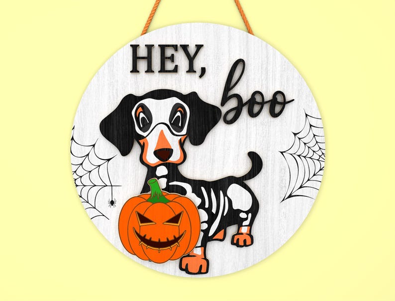 Może przedstawiać: Okrągły drewniany znak na Halloween z motywem szkieletu jamnika, pomarańczową dynią i napisem "HEY, boo". Znak ma białe tło z czarnymi pajęczynami i brązowym sznurkiem do zawieszenia.