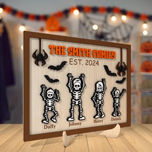 May include: A brown wooden sign with the text "The Smith Family Est. 2024" and five skeleton figures with names below them: "Duffy", "Johnny", "Henry", "Dennis". The sign has a Halloween theme with bats and spiders.