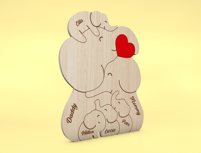 Op de afbeelding: Houten decoratie met olifantenfamilie thema, gegraveerde namen en een rood hart. De namen "Daddy", "Mommy", "Peter", "Carrie", "Hilton" en "Ellis" zijn gegraveerd op de olifantenfiguren. De decoratie is gemaakt van lichtgekleurd hout.