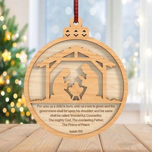 May include: Wooden Christmas ornament featuring a Nativity scene within a circular frame. The ornament includes a decorative top with a red and black plaid ribbon for hanging. The text from Isaiah 9:6 is printed on the ornament.