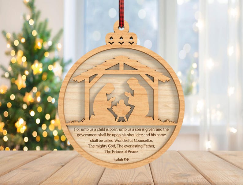 May include: Wooden Christmas ornament featuring a Nativity scene within a circular frame. The ornament includes a decorative top with a red and black plaid ribbon for hanging. The text from Isaiah 9:6 is printed on the ornament.