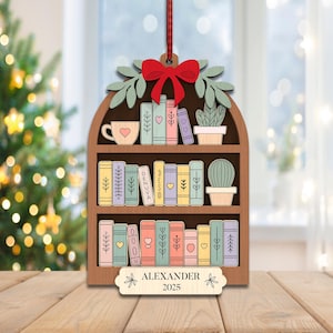 May include: Wooden bookshelf Christmas ornament with a red bow, green leaves, and a red and white striped ribbon. The ornament features a cup, a cactus, and colorful books. The name "ALEXANDER" and the year "2025" are displayed on the bottom.