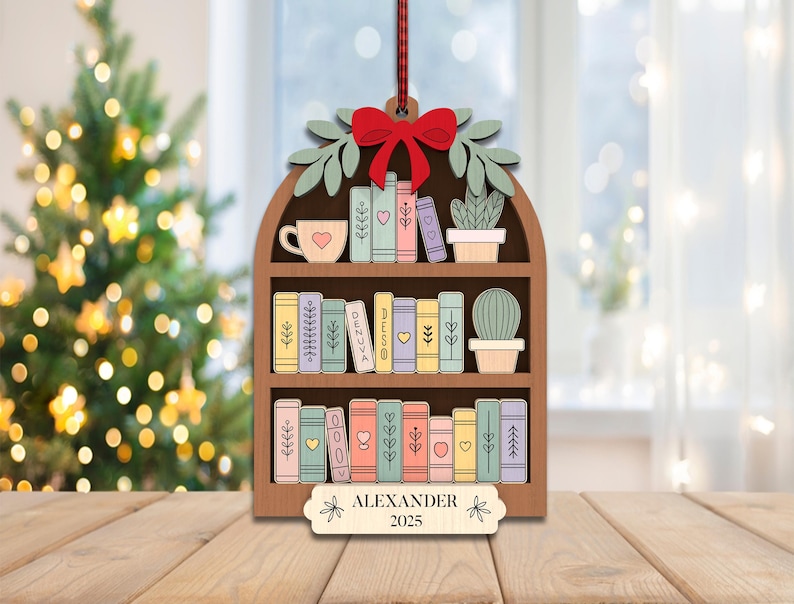 May include: Wooden bookshelf Christmas ornament with a red bow, green leaves, and a red and white striped ribbon. The ornament features a cup, a cactus, and colorful books. The name "ALEXANDER" and the year "2025" are displayed on the bottom.