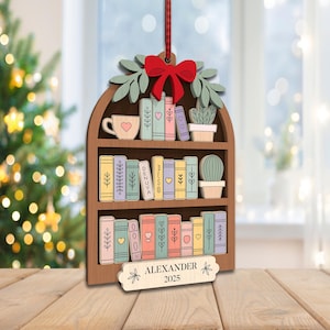 May include: Wooden Christmas tree ornament shaped like a bookshelf, with a red bow, green leaves, and a red ribbon. The bookshelf features colourful books, a teacup, and potted plants. The name "ALEXANDER" and the year "2025" are displayed on a small tag.