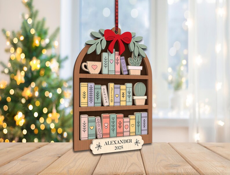 May include: Wooden Christmas tree ornament shaped like a bookshelf, with a red bow, green leaves, and a red ribbon. The bookshelf features colourful books, a teacup, and potted plants. The name "ALEXANDER" and the year "2025" are displayed on a small tag.