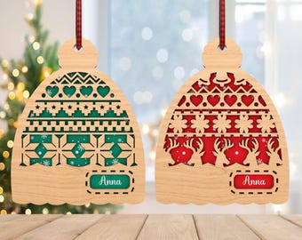 Könnte beinhalten: Zwei hölzerne Weihnachtsornamente in Form einer Wintermütze mit einem Schneeflocken- und geometrischen Muster. Die Ornamente sind rot und grün mit weißem Hintergrund und der Name "Anna" ist auf jedem Ornament geschrieben.