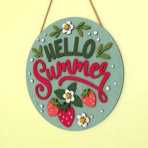 K&ouml;nnte beinhalten: Runder Holzschild mit den Worten "HELLO Summer" in gr&uuml;ner und roter Schrift. Das Schild zeigt Erdbeeren, Blumen und Bl&auml;tter. Es h&auml;ngt an einem braunen Seil vor einem hellgr&uuml;nen Hintergrund.