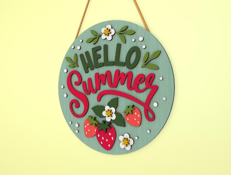 K&ouml;nnte beinhalten: Runder Holzschild mit den Worten "HELLO Summer" in gr&uuml;ner und roter Schrift. Das Schild zeigt Erdbeeren, Blumen und Bl&auml;tter. Es h&auml;ngt an einem braunen Seil vor einem hellgr&uuml;nen Hintergrund.
