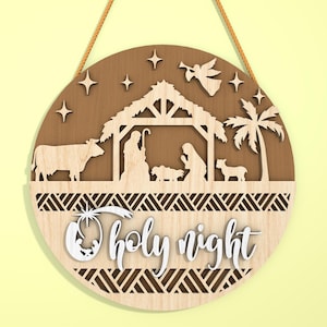 May include: A round wooden Christmas ornament with a nativity scene cut-out design. The ornament features a cow, angel, stars, and the words "holy night". It is suspended by a brown rope.