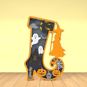 May include: A layered wooden Halloween decoration in the shape of the letter "J". The orange frame surrounds a spooky scene with ghosts, a haunted house, pumpkins, and a witch silhouette. A spider web and tombstone are also visible.