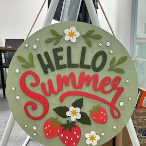 K&ouml;nnte beinhalten: Rundes Holzschild mit der Aufschrift "HELLO Summer" in roter Schrift. Das Schild ist hellgr&uuml;n mit roten Erdbeeren, wei&szlig;en Bl&uuml;ten und gr&uuml;nen Bl&auml;ttern. Es wird mit nat&uuml;rlichem Bindfaden aufgeh&auml;ngt.