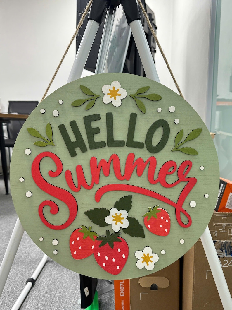 K&ouml;nnte beinhalten: Rundes Holzschild mit der Aufschrift "HELLO Summer" in roter Schrift. Das Schild ist hellgr&uuml;n mit roten Erdbeeren, wei&szlig;en Bl&uuml;ten und gr&uuml;nen Bl&auml;ttern. Es wird mit nat&uuml;rlichem Bindfaden aufgeh&auml;ngt.
