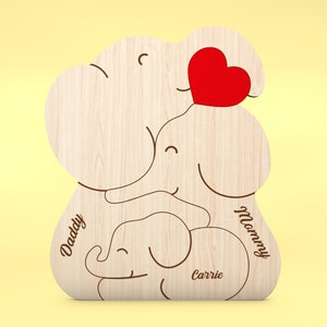 Op de afbeelding: Houten familie-olifantenbeeldje met een rood hart en de woorden "Daddy", "Mummy" en "Carrie" gegraveerd. Het beeldje is gemaakt van lichtgekleurd hout en heeft een gladde afwerking. De achtergrond is zachtgeel.