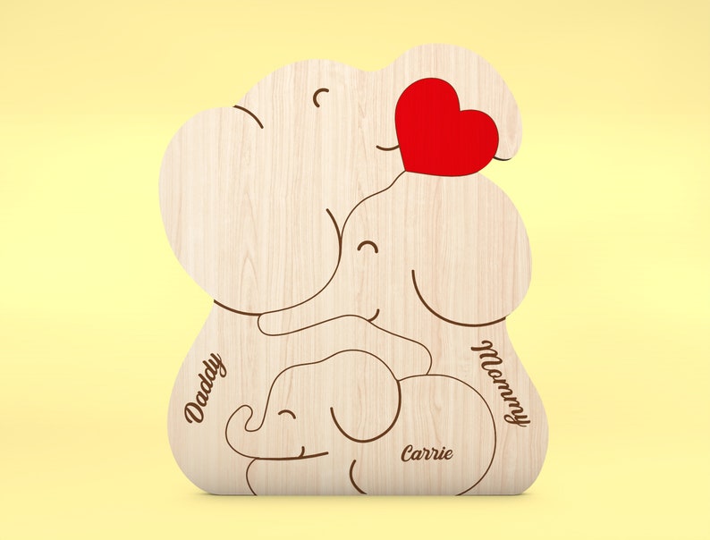 Op de afbeelding: Houten familie-olifantenbeeldje met een rood hart en de woorden "Daddy", "Mummy" en "Carrie" gegraveerd. Het beeldje is gemaakt van lichtgekleurd hout en heeft een gladde afwerking. De achtergrond is zachtgeel.