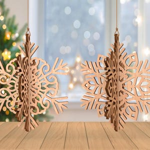 May include: Two wooden snowflake ornaments hanging from strings. The snowflakes have intricate cut-out designs and are made of light brown wood. A Christmas tree with twinkling lights is visible in the background.