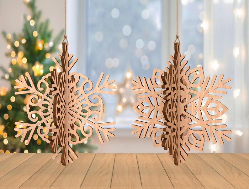 May include: Two wooden snowflake ornaments hanging from strings. The snowflakes have intricate cut-out designs and are made of light brown wood. A Christmas tree with twinkling lights is visible in the background.