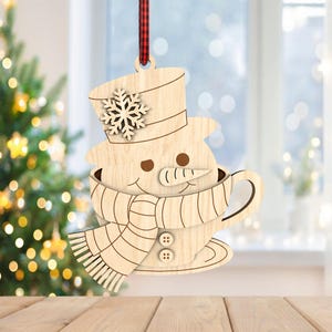 May include: Wooden snowman Christmas ornament, featuring a snowman in a teacup with a top hat and snowflake. The snowman has a carrot nose and button details on the scarf. A red and black plaid ribbon is attached for hanging. The ornament is displayed in front of a Christmas tree.