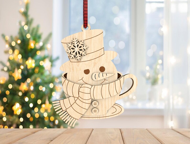 May include: Wooden snowman Christmas ornament, featuring a snowman in a teacup with a top hat and snowflake. The snowman has a carrot nose and button details on the scarf. A red and black plaid ribbon is attached for hanging. The ornament is displayed in front of a Christmas tree.