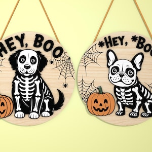 May include: Two round wooden Halloween signs with a dog and a French bulldog in skeleton costumes. Each sign has a jack-o'-lantern and spiderwebs. The signs read "HEY, BOO" in black letters and hang from a brown string.