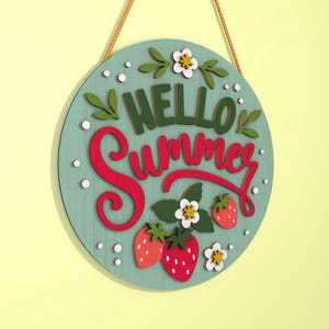 K&ouml;nnte beinhalten: Runder Holzschild mit den Worten "HELLO Summer" in gr&uuml;ner und roter Schrift. Das Schild zeigt Erdbeeren, wei&szlig;e Blumen und gr&uuml;ne Bl&auml;tter auf hellblauem Hintergrund. Ein braunes Seil ist zum Aufh&auml;ngen befestigt.
