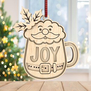 May include: Wooden Christmas ornament shaped like Santa Claus in a mug, with the word "JOY" cut out. The ornament has a handle and a belt design. It is hanging from a red and black plaid ribbon. A Christmas tree is in the background.