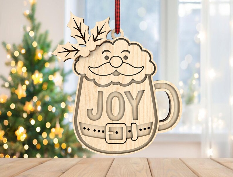 May include: Wooden Christmas ornament shaped like Santa Claus in a mug, with the word "JOY" cut out. The ornament has a handle and a belt design. It is hanging from a red and black plaid ribbon. A Christmas tree is in the background.