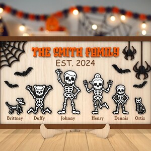 May include: A wooden sign with a Halloween theme featuring a family of skeletons. The sign reads "The Smith Family Est. 2024" and includes the names of each family member: Brittney, Duffy, Johnny, Henry, Dennis, and Ortiz.