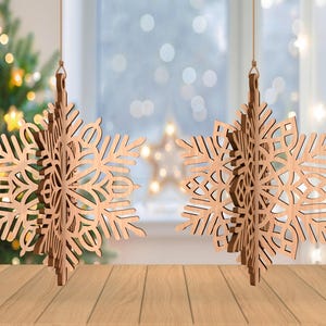 May include: Two wooden snowflake ornaments hanging from strings. The snowflakes have intricate cut-out designs and are made of light-colored wood. A Christmas tree with blurred lights is in the background, along with a window and a wooden surface.