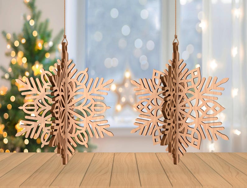 May include: Two wooden snowflake ornaments hanging from strings. The snowflakes have intricate cut-out designs and are made of light-colored wood. A Christmas tree with blurred lights is in the background, along with a window and a wooden surface.