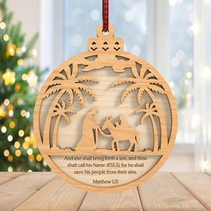 May include: Wooden Christmas ornament with a laser-cut nativity scene, featuring palm trees, figures, and text from Matthew 1:21. The ornament is round with a decorative top and a red and black plaid ribbon for hanging. A Christmas tree is in the background.