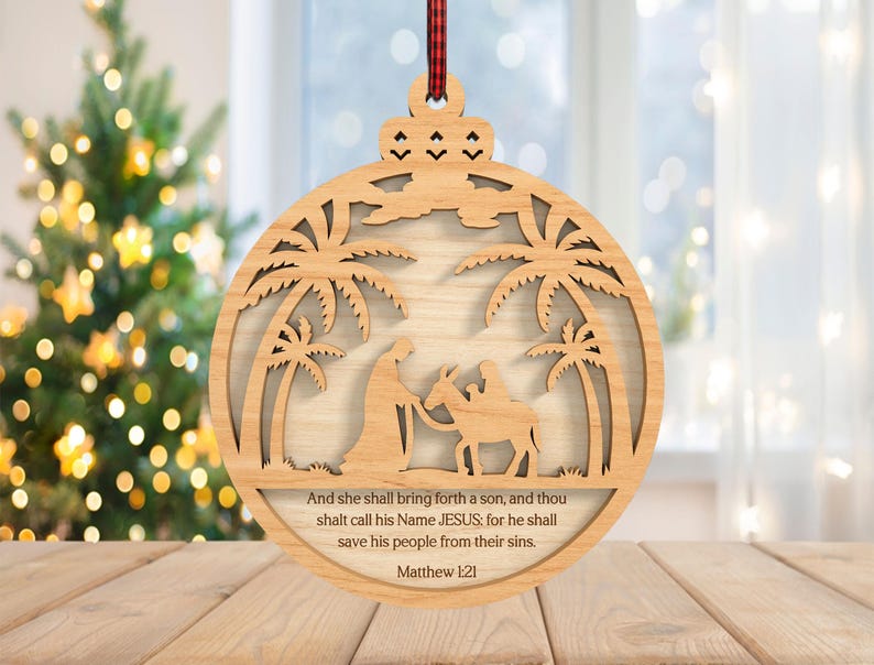 May include: Wooden Christmas ornament with a laser-cut nativity scene, featuring palm trees, figures, and text from Matthew 1:21. The ornament is round with a decorative top and a red and black plaid ribbon for hanging. A Christmas tree is in the background.