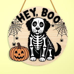 Może przedstawiać: Okrągły drewniany znak na Halloween z motywem szkieletu psa, dynią i napisem "HEY, BOO". Znak ma jutową linę do zawieszenia i zawiera pajęczyny i pająki.