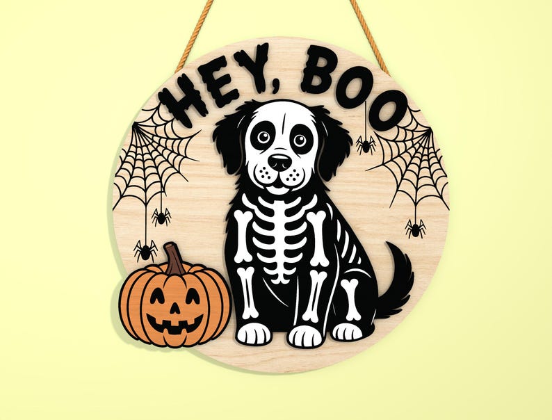 Może przedstawiać: Okrągły drewniany znak na Halloween z motywem szkieletu psa, dynią i napisem "HEY, BOO". Znak ma jutową linę do zawieszenia i zawiera pajęczyny i pająki.