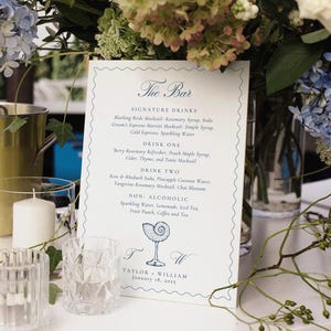 May include: A wedding bar display featuring a menu, flowers, and glasses. The menu details signature and non-alcoholic drinks. A gold ice bucket and candles enhance the setting. The menu reads "The Bar".