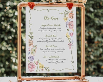 May include: A copper-colored metal frame holds a white sign with the text "the Bar" and a list of drink options. Floral illustrations and dried flowers adorn the frame. The sign lists signature drinks, including pomegranate spritzer and non-alcoholic options.