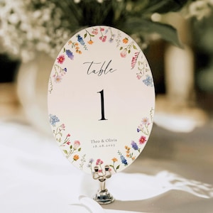 Puede incluir: Una tarjeta ovalada de número de mesa con un borde floral de flores silvestres coloridas. La tarjeta muestra el texto Table 1, seguido de los nombres Theo & Olivia y la fecha 18.08.2023. La tarjeta se sostiene verticalmente con un soporte de metal plateado.