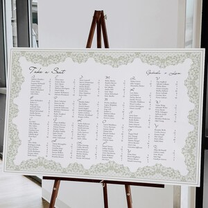 May include: A large white seating chart with a decorative border, displayed on a wooden easel. The chart features names arranged in columns, with the words "Take a Seat" and names at the top. The overall design is elegant and suitable for a wedding or event.