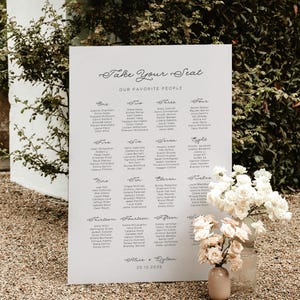 May include: A large white wedding seating chart with elegant calligraphy that reads "Take Your Seat" and "Our Favorite People." The chart lists names in numbered sections. A vase of white flowers sits in front of the chart.